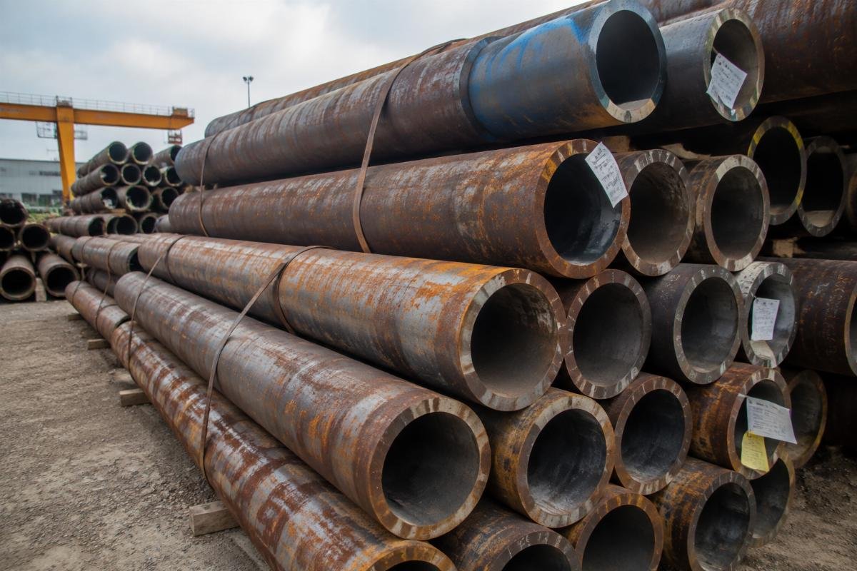 Stacked structural steel pipes in an open yard showing signs of surface corrosion and mixed labeling